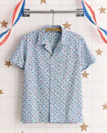 The Micro Garden Shirt - S features a light blue, short-sleeve design with red and blue floral prints, made from a unique textile inspired by 1930s-1940s fabrics. Displayed on a hanger with festive decor in the background.