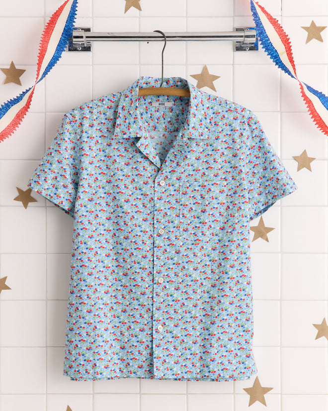 The Micro Garden Shirt - S features a light blue, short-sleeve design with red and blue floral prints, made from a unique textile inspired by 1930s-1940s fabrics. Displayed on a hanger with festive decor in the background.