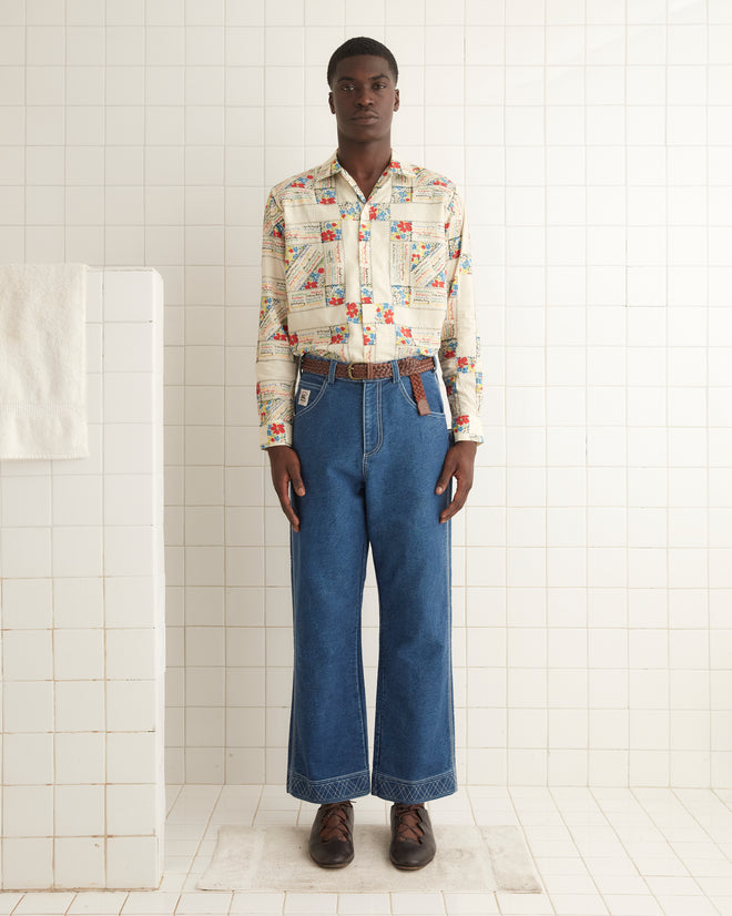 A person stands in a tiled room wearing a White Micro Signature Long Sleeve Shirt with intricate patterns, blue jeans featuring antique patchwork near the hem, and brown shoes.