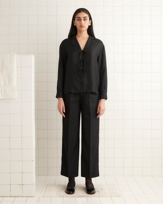 A person in a Micro Tie Long Sleeve Shirt - Black stands against a tiled white wall, showcasing a minimalist setting.