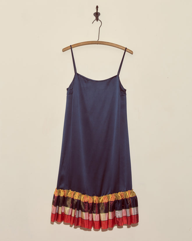 The Midnight Gala Dress - Navy is a silk satin dress with thin straps and a colorful ruffled hem, hanging on a wooden hanger—a unique piece that brings distinctive charm to any wardrobe.