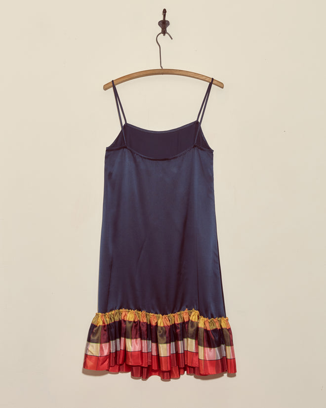 The Midnight Gala Dress - Navy is a unique silk satin slip dress with thin straps and a vibrant ruffled hem, displayed elegantly on a wooden hanger.