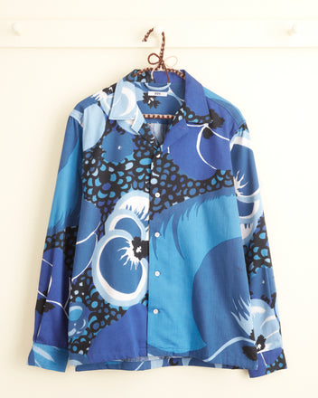 The Mink Protea Long Sleeve Shirt in size S/M, known for its unique swirling blue floral pattern, is hanging on a white wall.