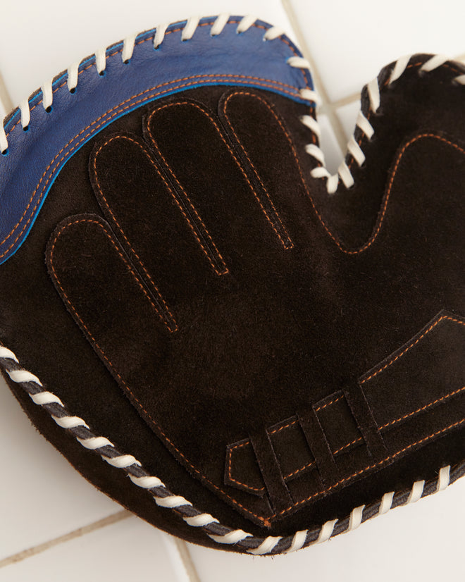 The Mitt Clutch, featuring a sleek black and blue design with white stitching, elegantly rests on a white tiled surface, adding a touch of sophistication to any sports enthusiast's collection.