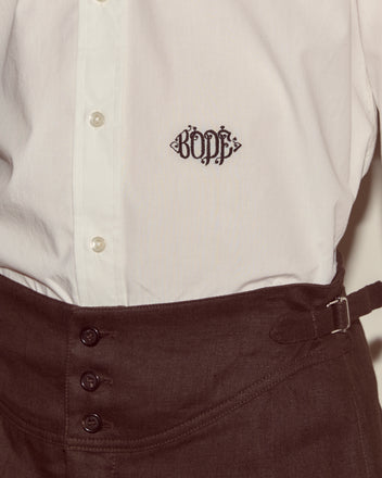 A Monogrammed Poplin Shirt in white, made from 100% cotton with "BODE" embroidered on the chest, styled with high-waisted brown pants.