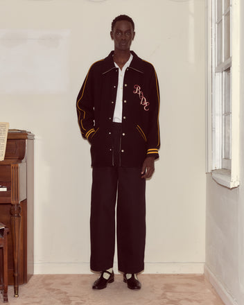 A person in black pants and a Monogram Snap Reversible Jacket stands indoors by a piano and a window.