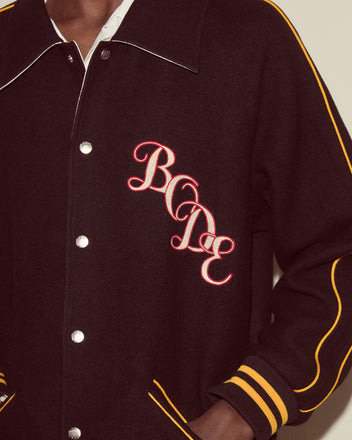 Person wearing the Monogram Snap Reversible Jacket, a black reversible jacket with yellow stripes and a red-and-white chain-stitched Bode logo on the chest.