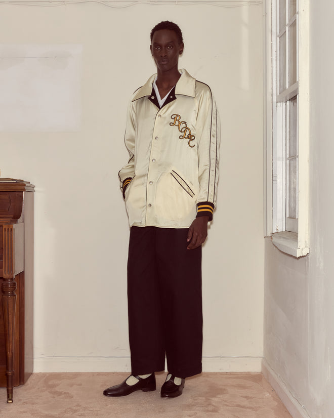 A person in a cream Monogram Snap Reversible Jacket with a chain-stitched Bode logo and wide black pants stands indoors by a window, hands in pockets.