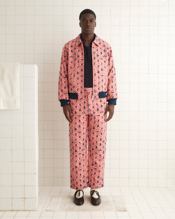 A person stands in a white-tiled room wearing the Monte Rosa Jacket, a 1960s-inspired pink patterned piece, paired with matching pants, a dark shirt underneath, and black shoes.