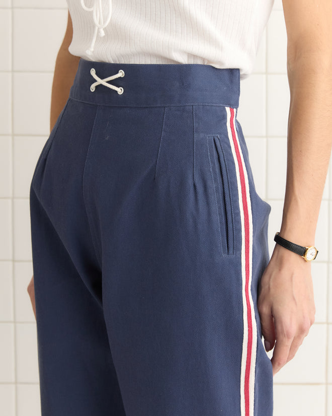 A person stands against a tiled wall, wearing Monterrey Stripe Trousers in heavyweight blue twill with red and white stripes, paired with a white top.