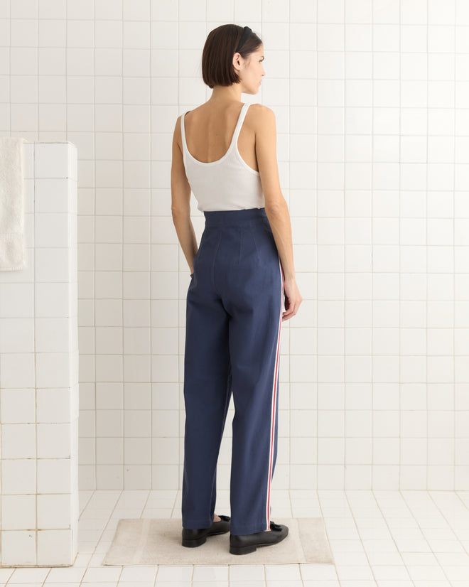 A woman in a white tank top and Monterrey Stripe Trousers, featuring a 1930s style with a drawstring waist, stands on a mat in a tiled room, facing away.