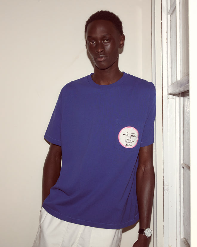 A person wearing the Moon Tee, featuring an embroidered face design, stands by a white wall and window paired with white pants.