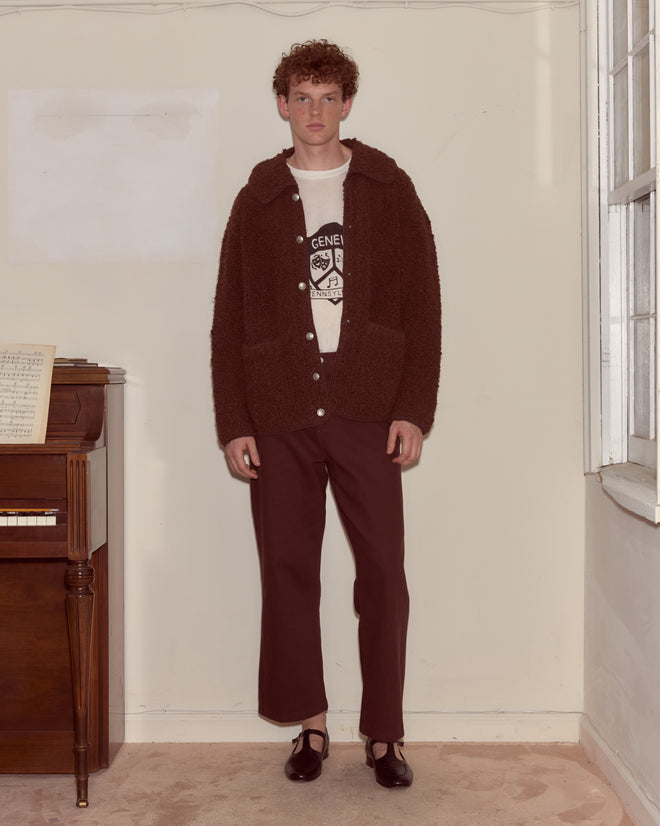 A man wearing the Moppet Cardigan - Brown, featuring a textured design and oversized silhouette, stands indoors by a piano near a window, looking at the camera.