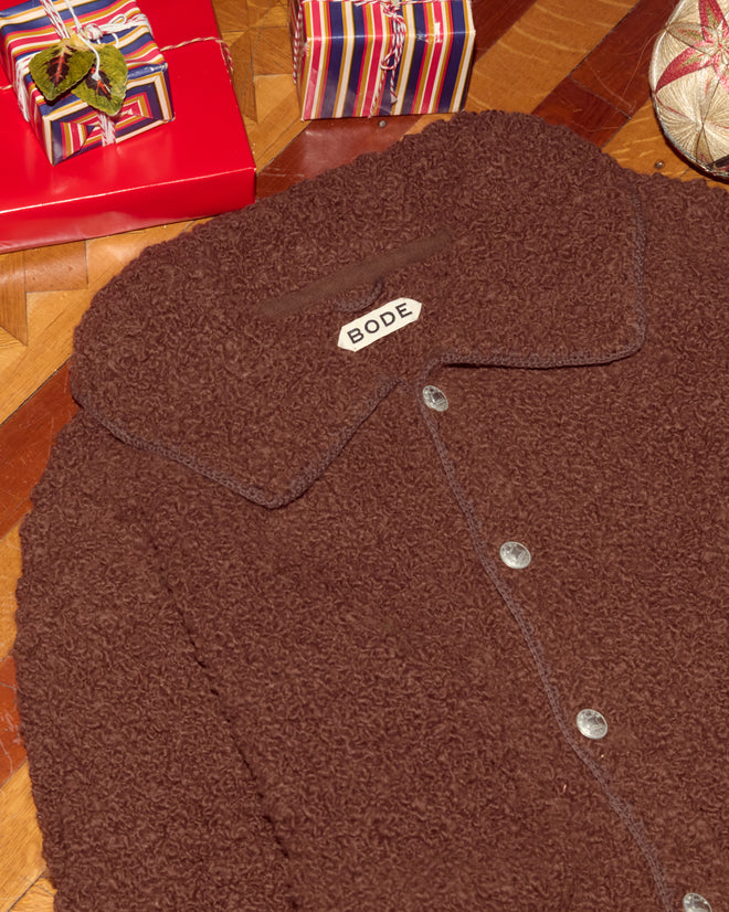 A brown Moppet Cardigan from BODE with a textured finish and oversized fit lies near wrapped gifts on a wooden floor.