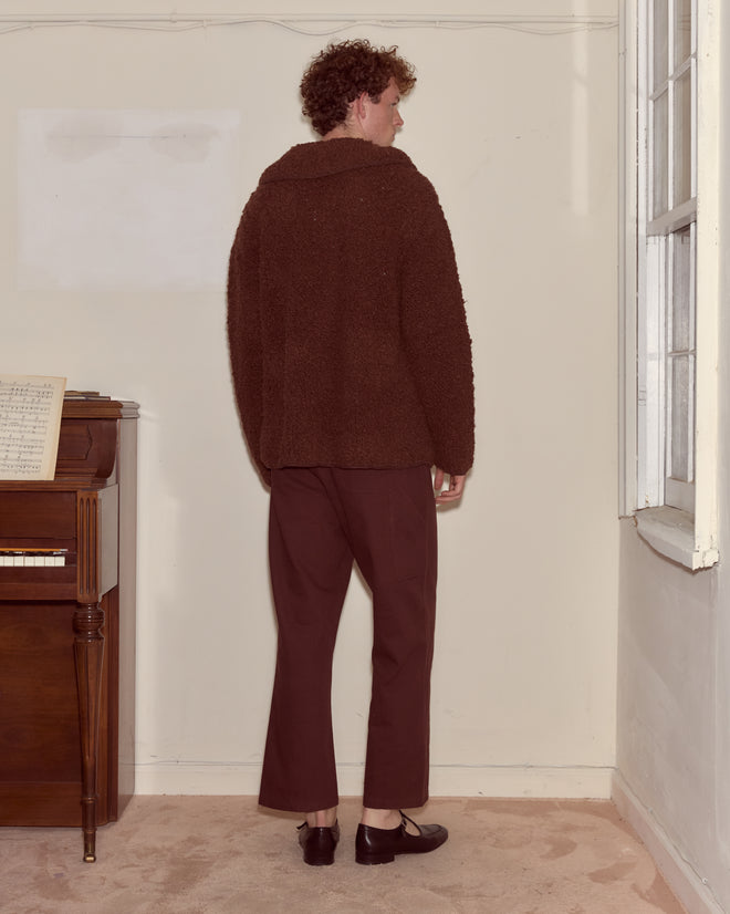 A person with curly hair wears the Moppet Cardigan - Brown indoors, standing by a piano and window, facing away.