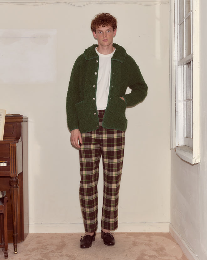 A person in a Moppet Cardigan - Green stands indoors by a piano and a window, wearing plaid pants.