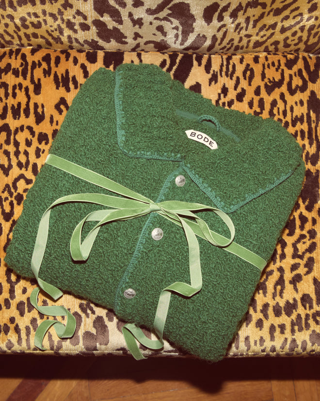 The Moppet Cardigan - Green, with ribbon and buttons, is folded on leopard-print fabric, evoking the cozy texture of a bouclé-knit cardigan.