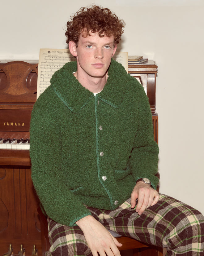 A young person with curly hair wears the oversized Moppet Cardigan in green while sitting by a Yamaha piano, sheet music visible behind them.