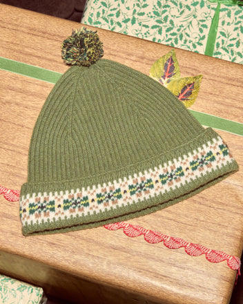 The Moritz Hat, a green Fair Isle knit with pom-pom, made from soft merino wool, rests on a wooden surface beside wrapped gifts—ideal for fans of luxury accessories.