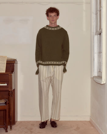 Indoors by a piano and window, a person wears the Moritz Sweater, crafted in green virgin wool, paired with white striped pants.
