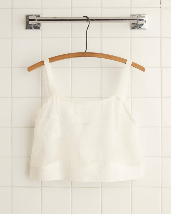 The Morning Lilies Tank - XS, a unique white spaghetti strap crop top adorned with vintage floral appliqués, hangs on a wooden hanger against a tiled wall.