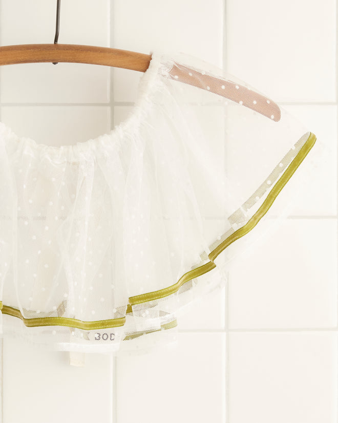 A white sheer ruffled garment with green trim, named the Moss Velvet Collar and reminiscent of 1950s costumes, is hung on a twisted brown hanger against a light wall.