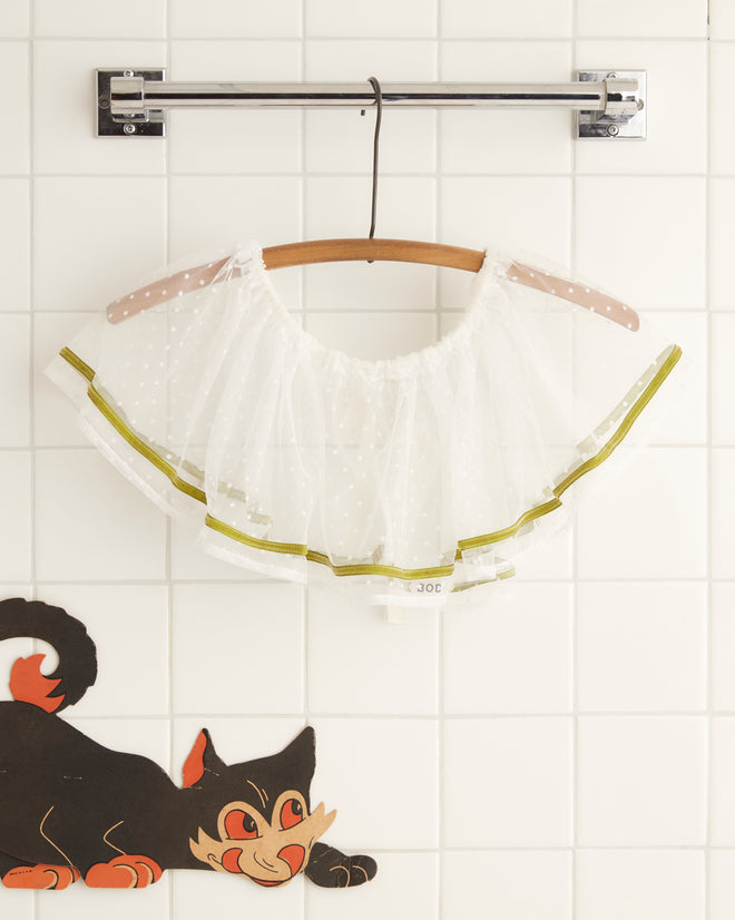 A white sheer ruffled garment with green trim, named the Moss Velvet Collar and reminiscent of 1950s costumes, is hung on a twisted brown hanger against a light wall.