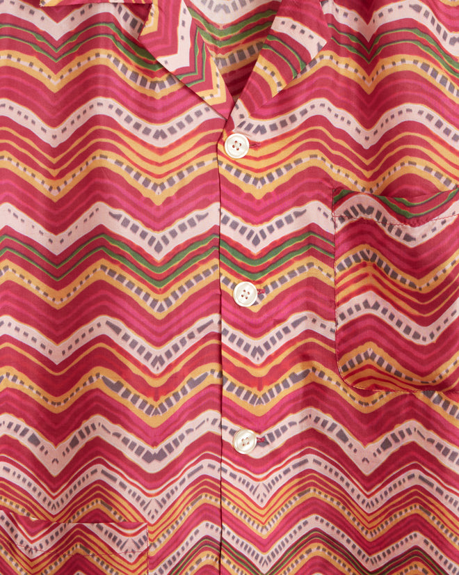 The Mothra Zig-Zag Short Sleeve Shirt, made from 100% silk and featuring a vibrant pink, red, and orange zigzag pattern created through traditional Rajasthani Mothra dyeing, hangs on a wall hook.