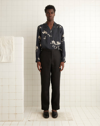 A person stands in a white tiled room, wearing a dark floral shirt and the Linen Suiting Trousers in black.