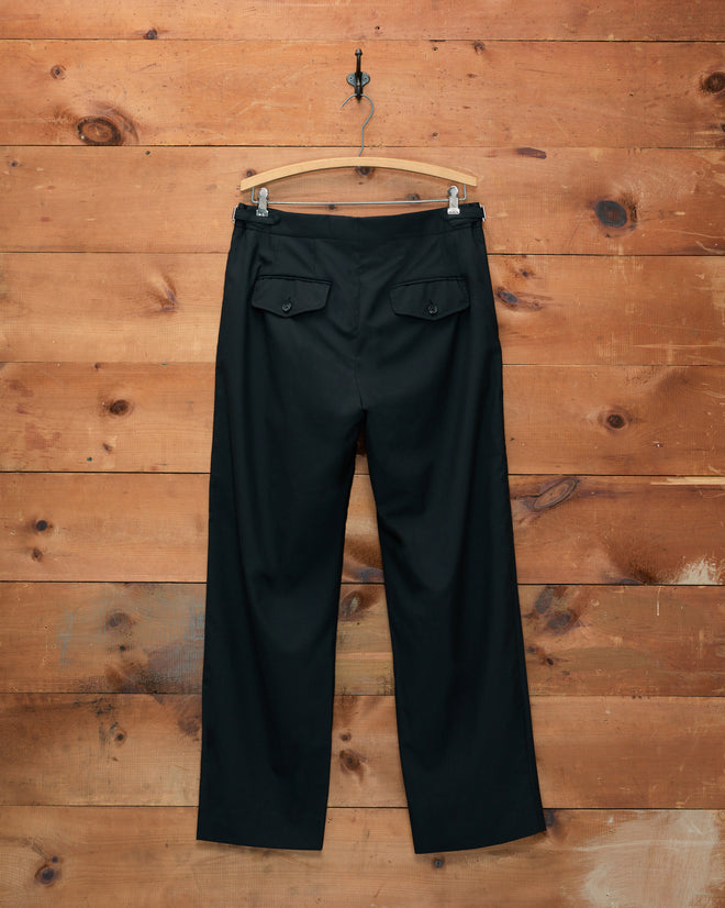 Tropical Wool Side-Buckle Trousers - Black with a straight-leg silhouette and flap back pockets are displayed on a hanger against a wooden wall. Crafted from lightweight tropical wool, these midcentury-inspired trousers offer timeless sophistication and comfort.