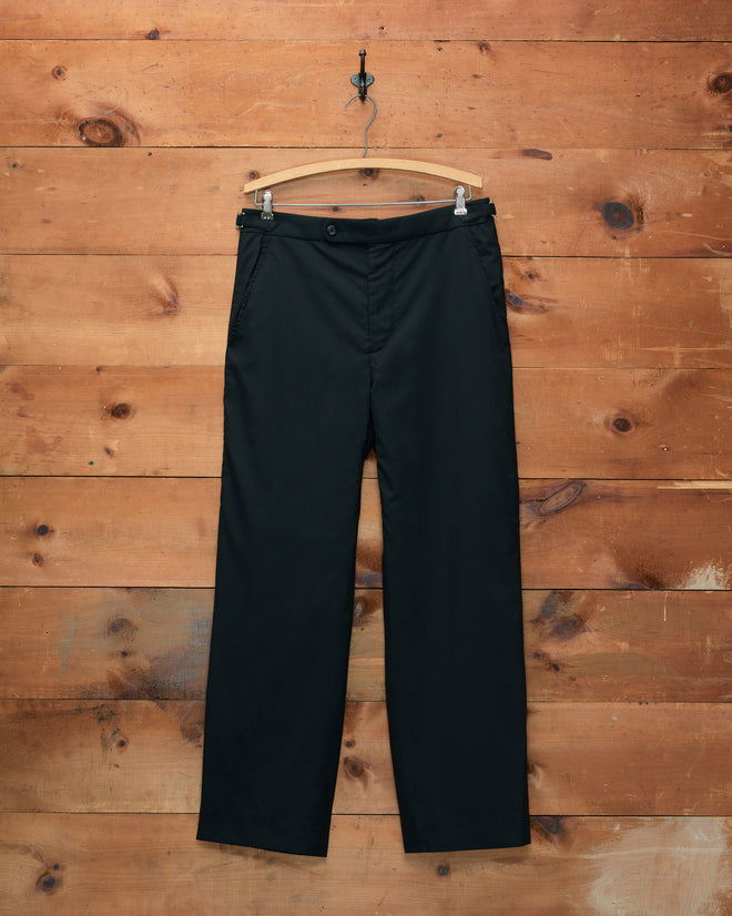 Tropical Wool Side-Buckle Trousers - Black, displayed on a wooden hanger against a rustic wooden wall.