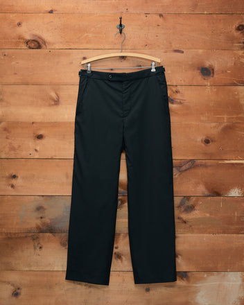 Tropical Wool Side-Buckle Trousers - Black, displayed on a wooden hanger against a rustic wooden wall.