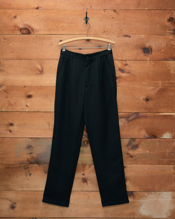 Tropical Wool Double-Button Trousers hang elegantly on a wooden hanger against a rustic wooden wall background. Crafted from lightweight wool, these midcentury suiting trousers blend timeless sophistication with modern comfort.