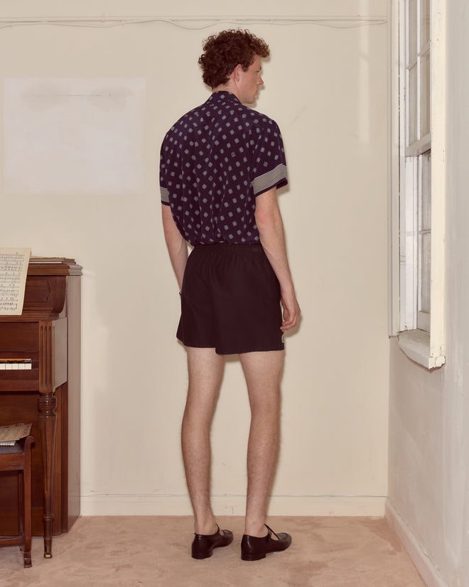 Man standing in a corner facing the wall, wearing a patterned shirt, Field Shorts - Black, and dress shoes.