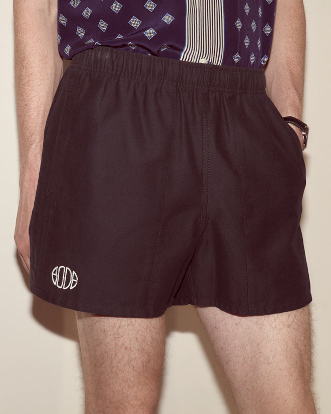 A person wearing Field Shorts - Black, made from dark heavyweight cotton twill with the BODE logo, paired with a patterned shirt and hands in pockets.