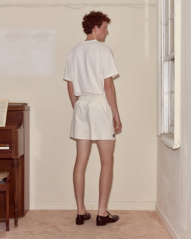 A man in a heavyweight cotton twill white T-shirt and Field Shorts - White stands facing a wall in a sparsely furnished room with a piano.
