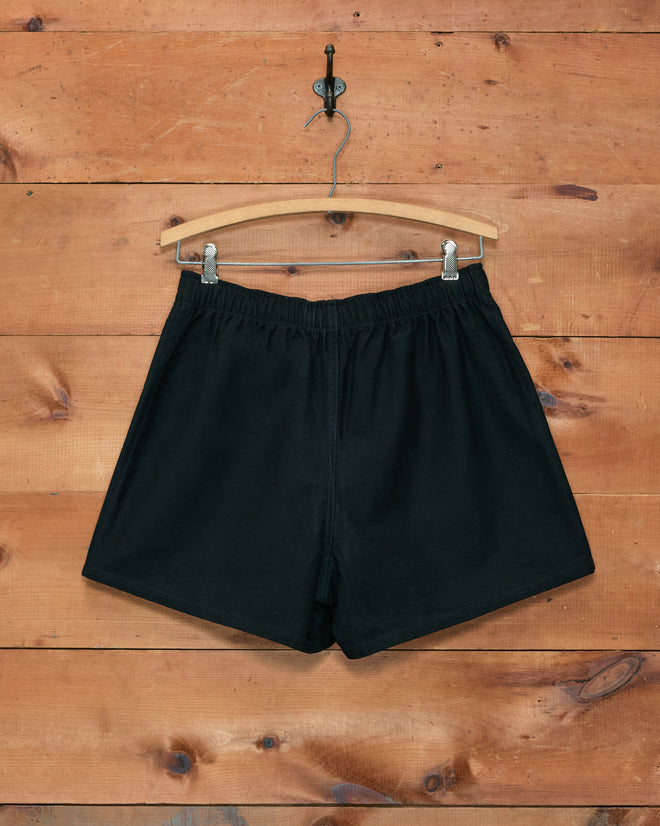 Field Shorts - Black, made from heavyweight cotton twill and inspired by the iconic 1980s rugby short silhouette, displayed on a wooden hanger against a wooden plank wall background.