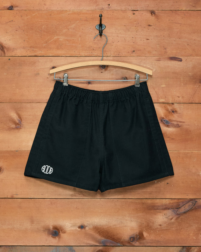 Field Shorts - Black from Bode, inspired by 1980s rugby styles, featuring a white Bode logo and displayed on a wooden hanger against a wooden wall. Expertly crafted from heavyweight cotton twill for exceptional durability and classic appeal.
