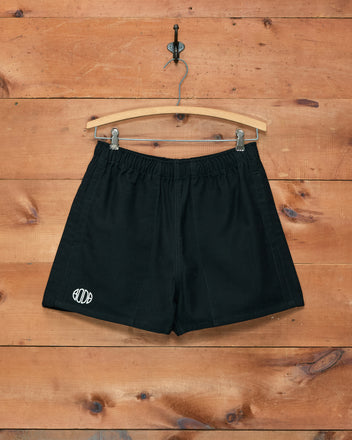 Field Shorts - Black from Bode, inspired by 1980s rugby styles, featuring a white Bode logo and displayed on a wooden hanger against a wooden wall. Expertly crafted from heavyweight cotton twill for exceptional durability and classic appeal.