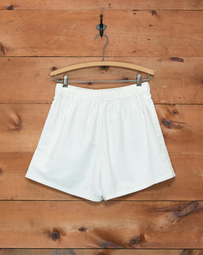 Field Shorts - White, crafted from heavyweight cotton twill and inspired by 1980s rugby shorts, are displayed on a wooden hanger against a wooden wall background. These shorts feature an embroidered Bode logo for a distinctive touch.