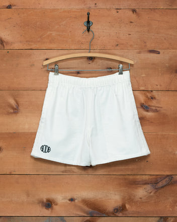 Field Shorts - White, inspired by 1980s rugby shorts and featuring a small black logo, hang on a wooden hanger against a wooden wall.
