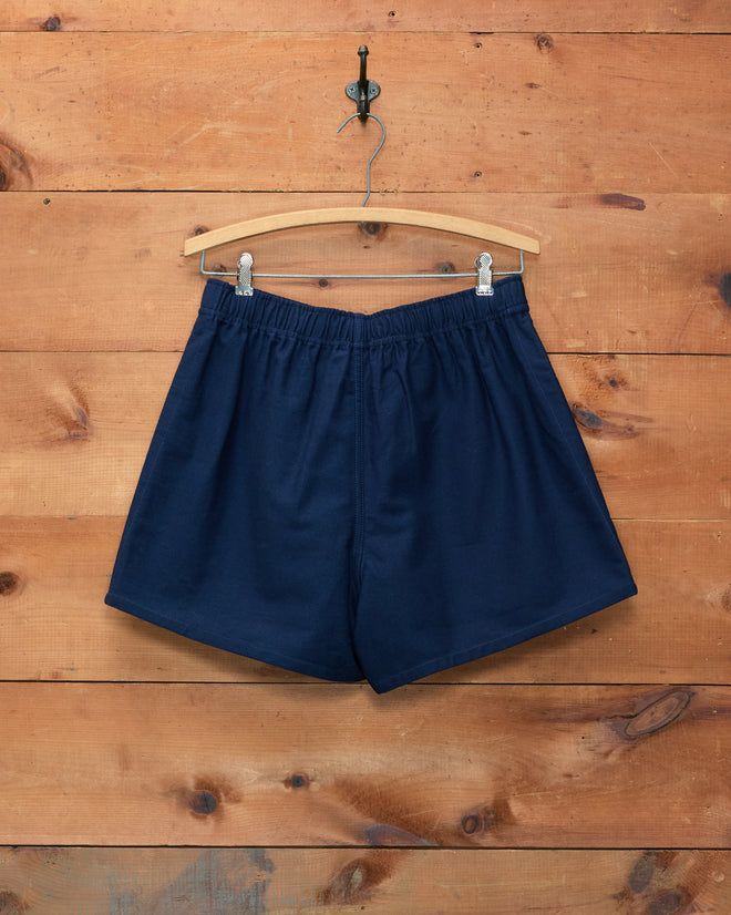 Field Shorts - Navy, crafted from heavyweight cotton, are displayed on a wooden hanger against a wooden wall background and finished with the distinctive Bode logo for a classic touch.