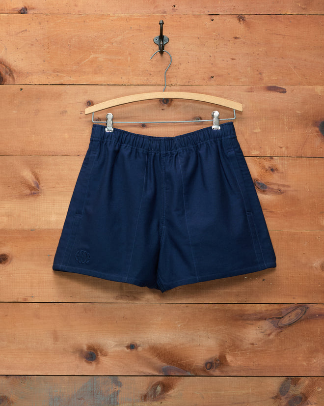 Field Shorts - Navy, crafted from heavyweight cotton, hang on a wooden hanger against a wooden wall background and subtly display the signature Bode logo.