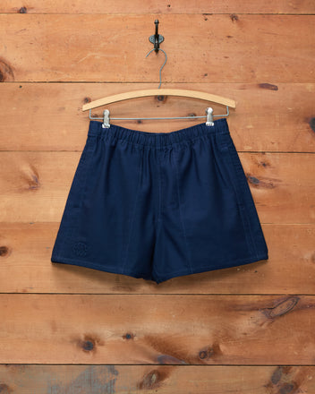 Field Shorts - Navy, crafted from heavyweight cotton, hang on a wooden hanger against a wooden wall background and subtly display the signature Bode logo.