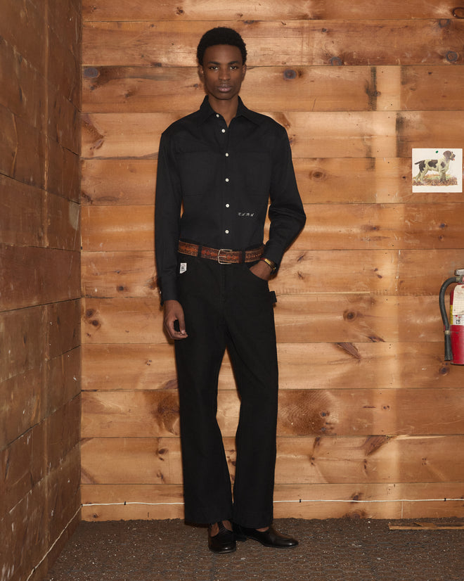 A person wearing a black shirt and Embroidered Denim Knolly Brook Trousers - Black stands against wooden walls near a fire extinguisher and a small dog photo.