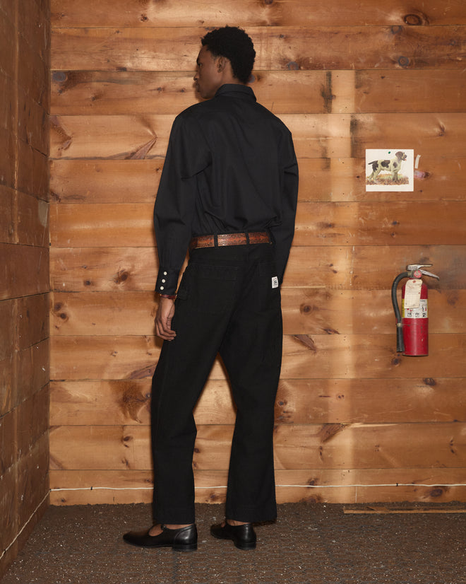 Person in black shirt and Embroidered Denim Knolly Brook Trousers - Black stands near a wooden wall with fire extinguisher and small photo on the wall.