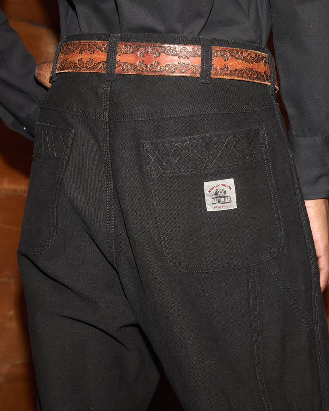 Person wearing Embroidered Denim Knolly Brook Trousers in black, shown from behind, featuring back pockets and a brown patterned belt.