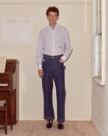A man in a striped shirt and Embroidered Denim Knolly Brook Trousers stands indoors beside a piano and a window.