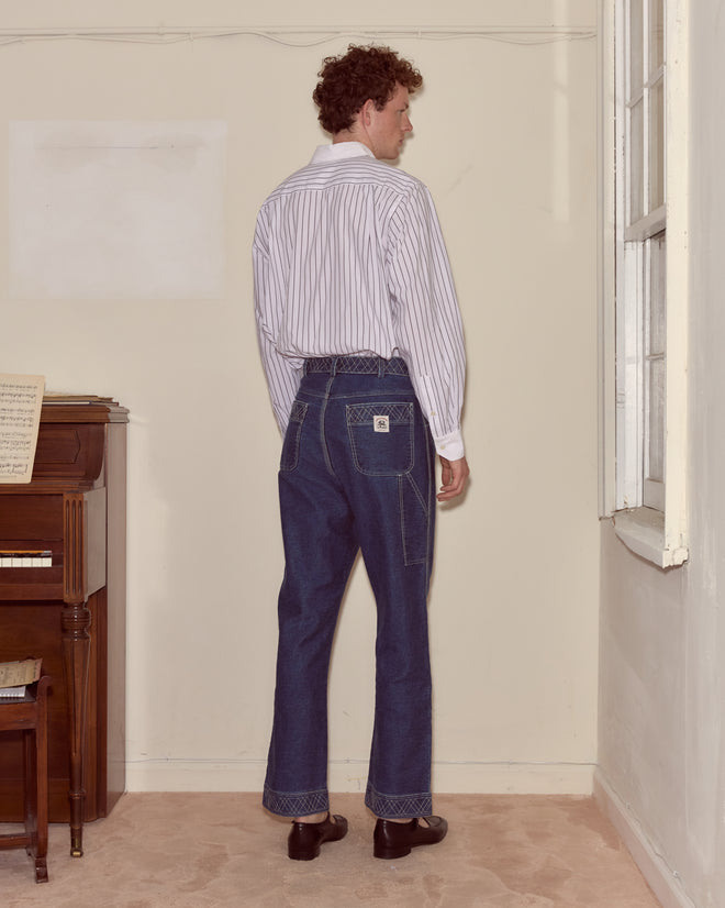 A man with curly hair in a striped shirt and Embroidered Denim Knolly Brook Trousers stands facing a bare wall near a piano and window.
