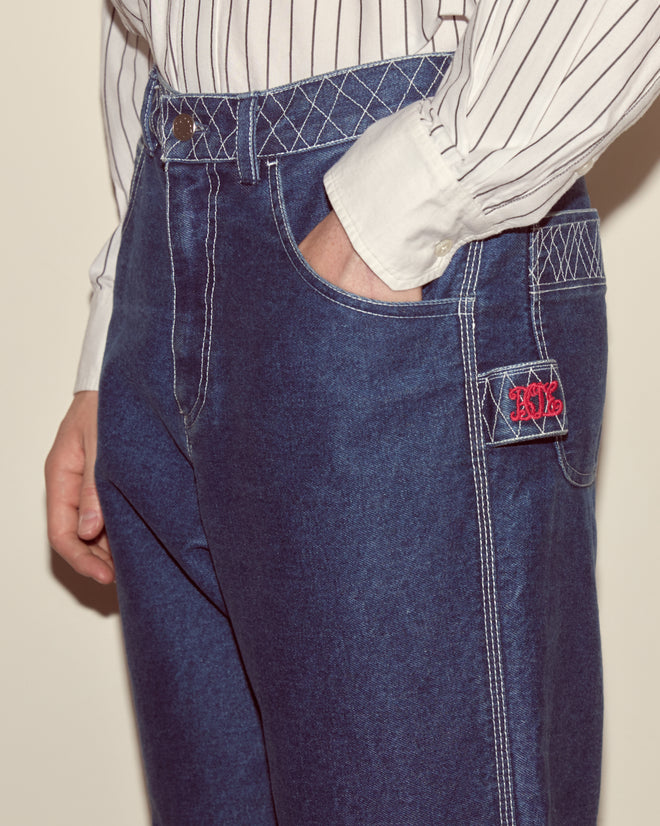 Wearing Embroidered Denim Knolly Brook Trousers—100% cotton with white stitching, red NY patch, and a striped shirt, hand in pocket.